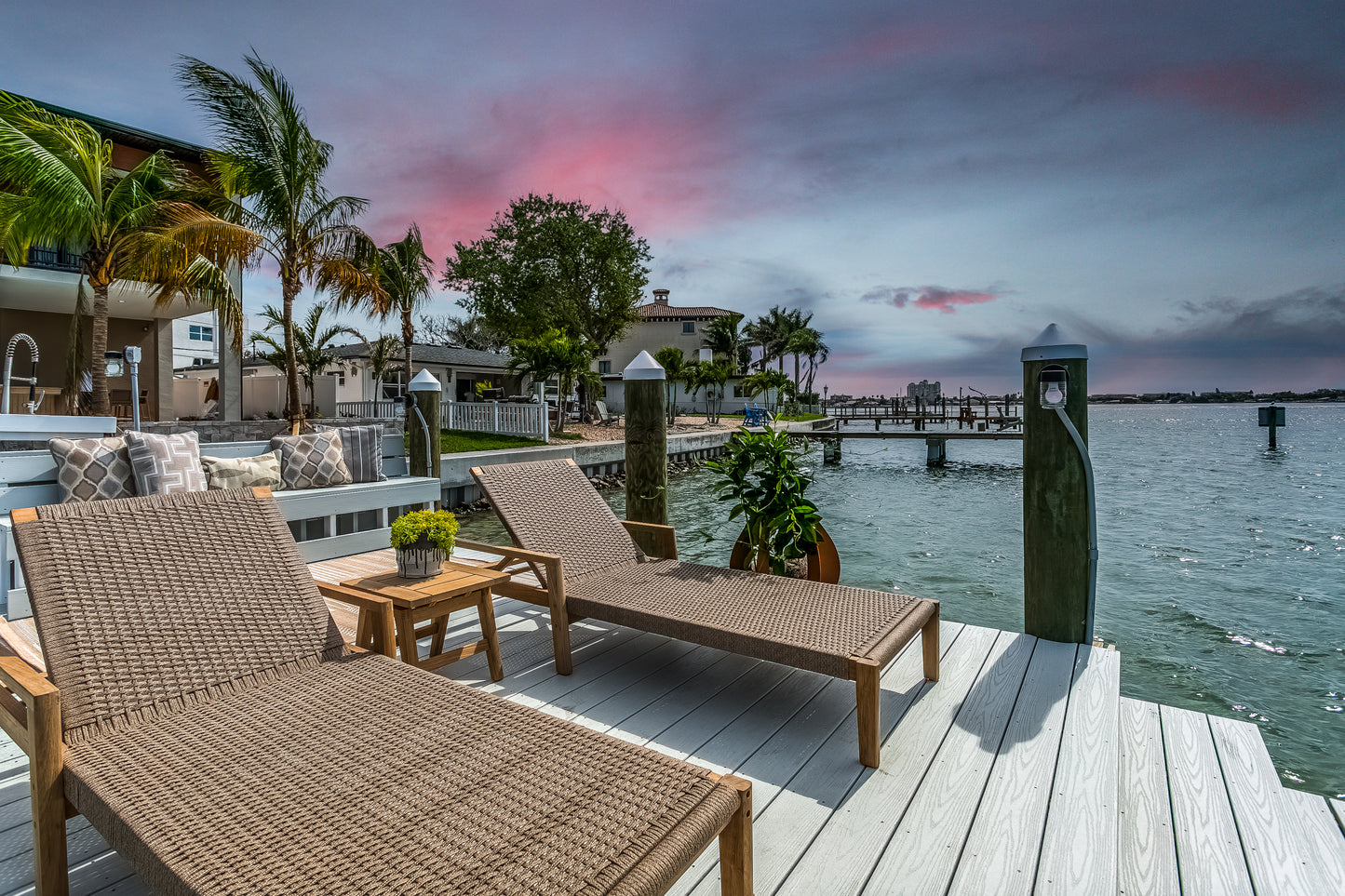 Two wicker chairs on a dock by a waterfront with palm trees and a colorful sky.