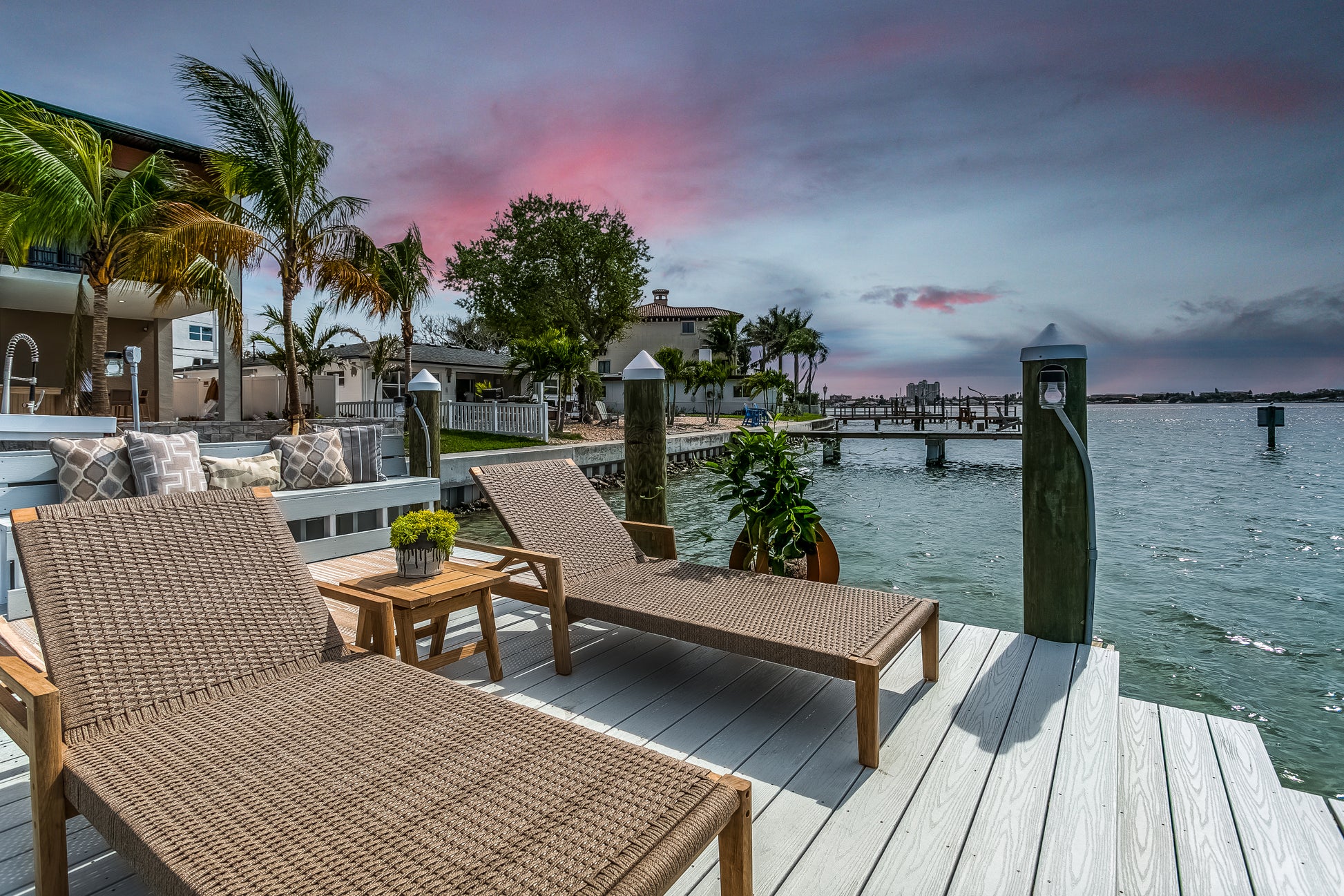 Two wicker chairs on a dock by a waterfront with palm trees and a colorful sky.