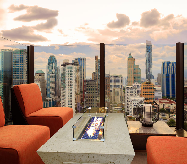 Outdoor seating area with a fire pit table and orange chairs, overlooking a city skyline.