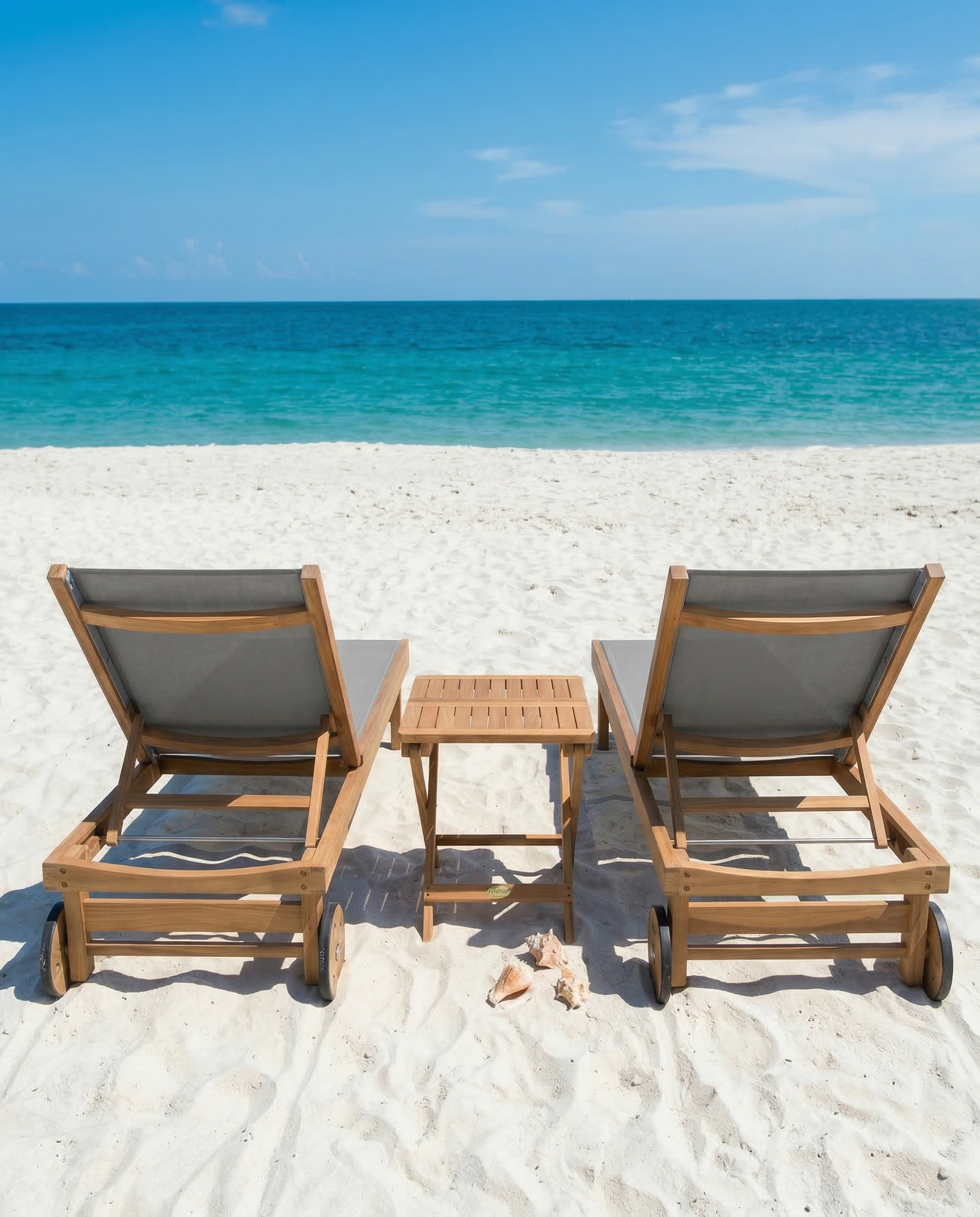 Two wooden beach chairs with a small table on a sandy beach with clear blue water and sky.