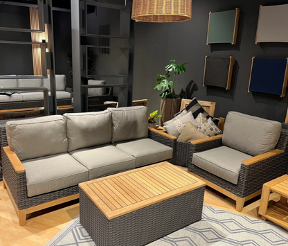 A three-seater sofa with beige cushions and a wooden frame, displayed in a showroom setting with decorative items and a large painting in the background.
